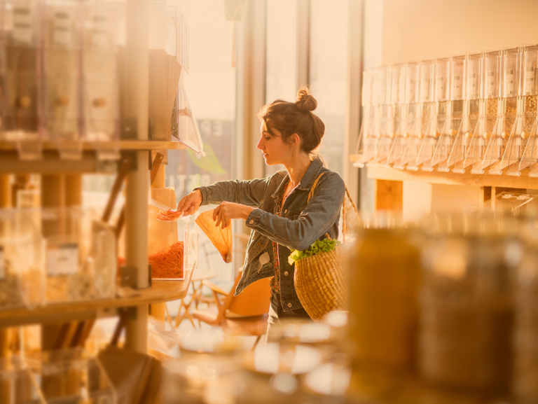 Junge Frau, mit großem Bast-Shopper über der Schulter, bedient sich im Unverpackt-Laden mit Schäufelchen und Papiertüte an einem großen Spender