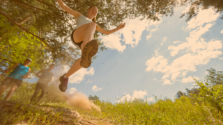 Junge Sportlerin wird beim Sprung über einen Graben von unten fotografiert, dabei fliegt der Dreck um das Objektiv