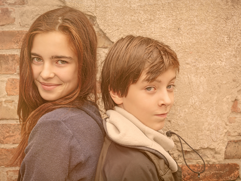 Geschwister stehen Rücken an Rücken lächelnd vor einer Hausmauer