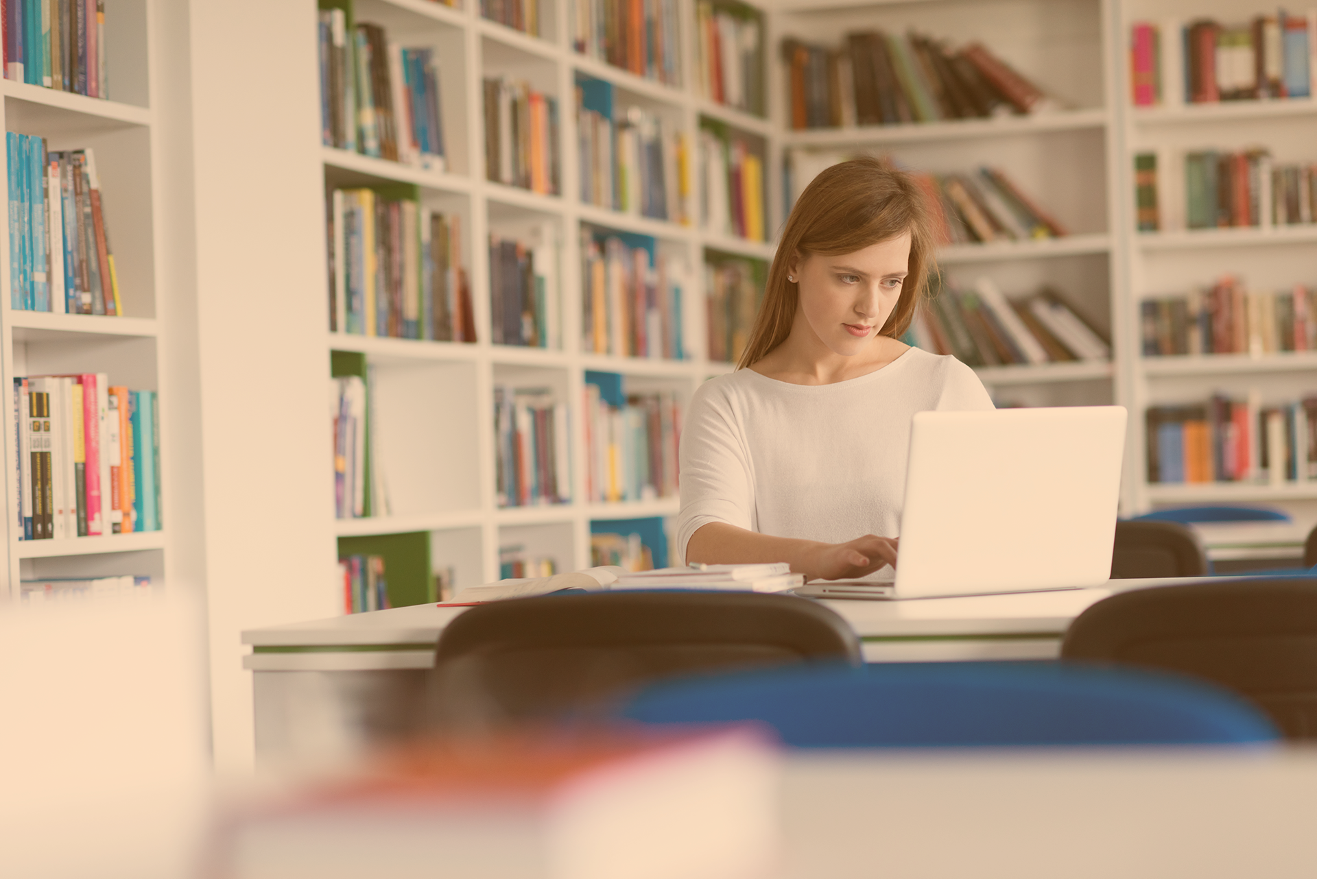 Jugendliche sitzt in einem hellen Raum vor Bücherregalen schreibend am Laptop, um sie herum liegen Unterlagen auf dem Tisch