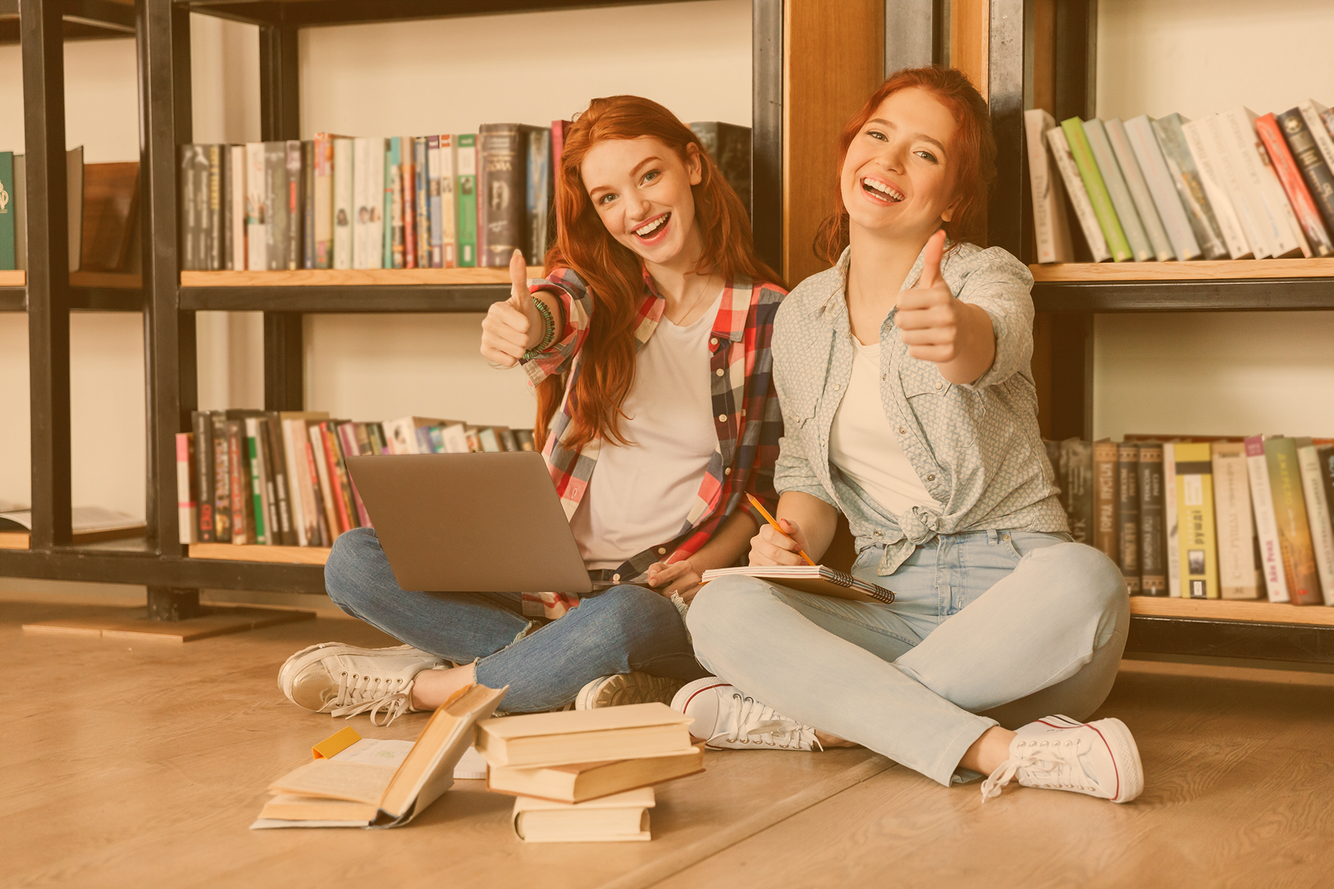 Zwei Jugendliche sitzen vor einem Bücherregal auf dem Boden und halten eine Daumen hoch