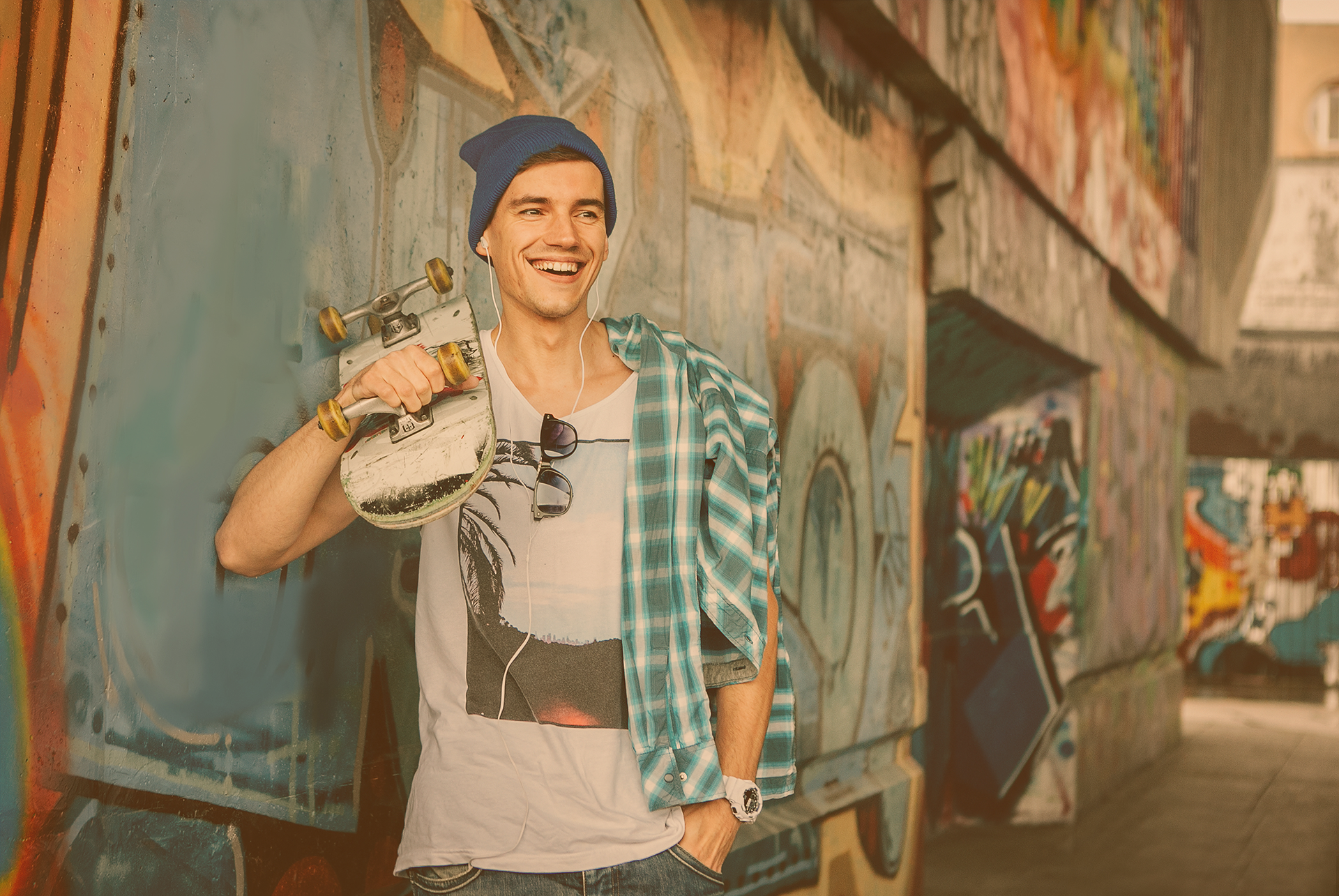 Junger Skater mit Hippster-Mütze steht mit Skateboard auf der Schulter an einer Graffiti-Wand gelehnt