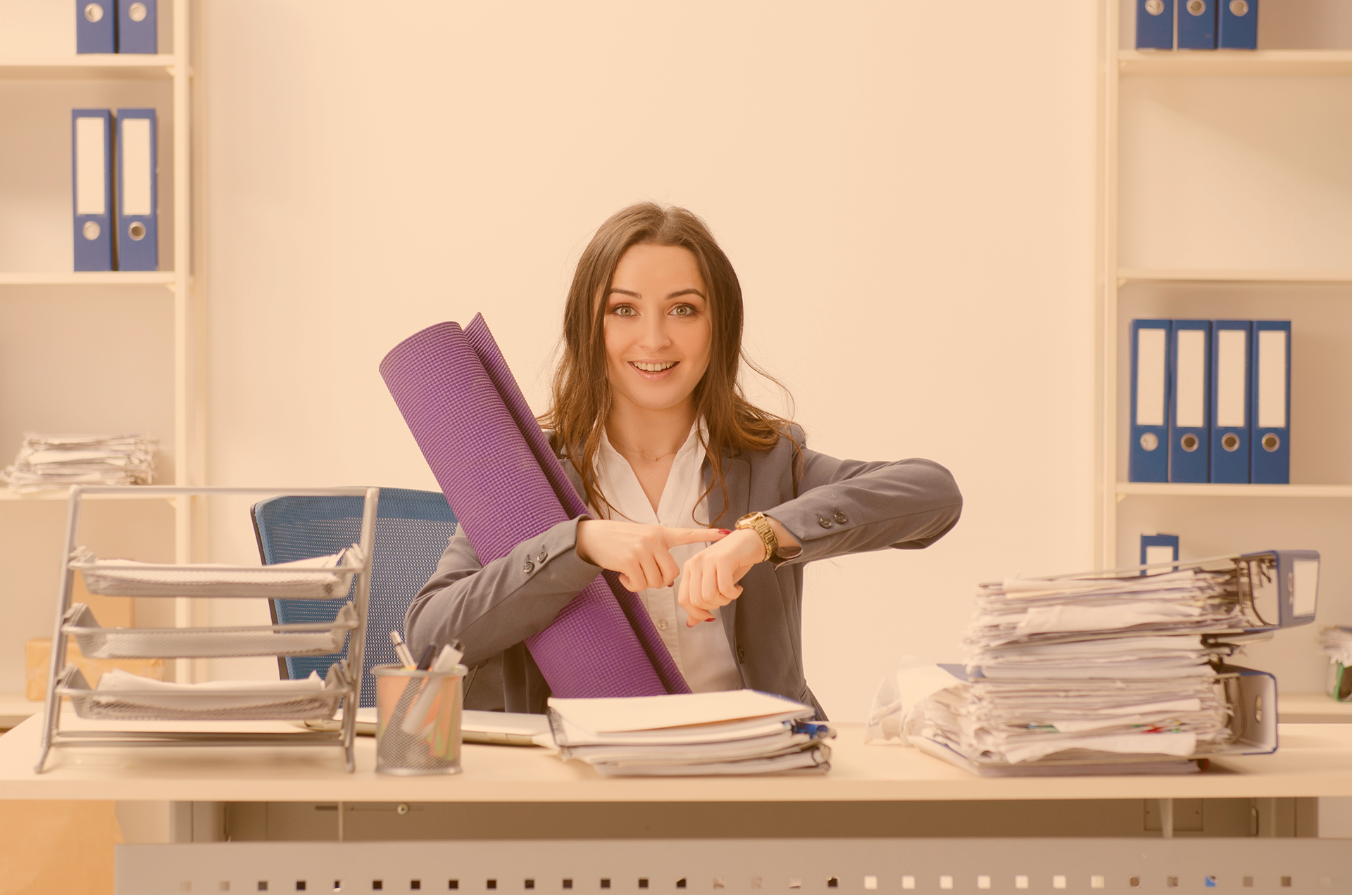 Junge Frau sitzet hinter vollem Schreibtisch und zeigt auf ihre Armbanduhr, und trägt unter dem Arm eine gerollte Gymnastikmatte 