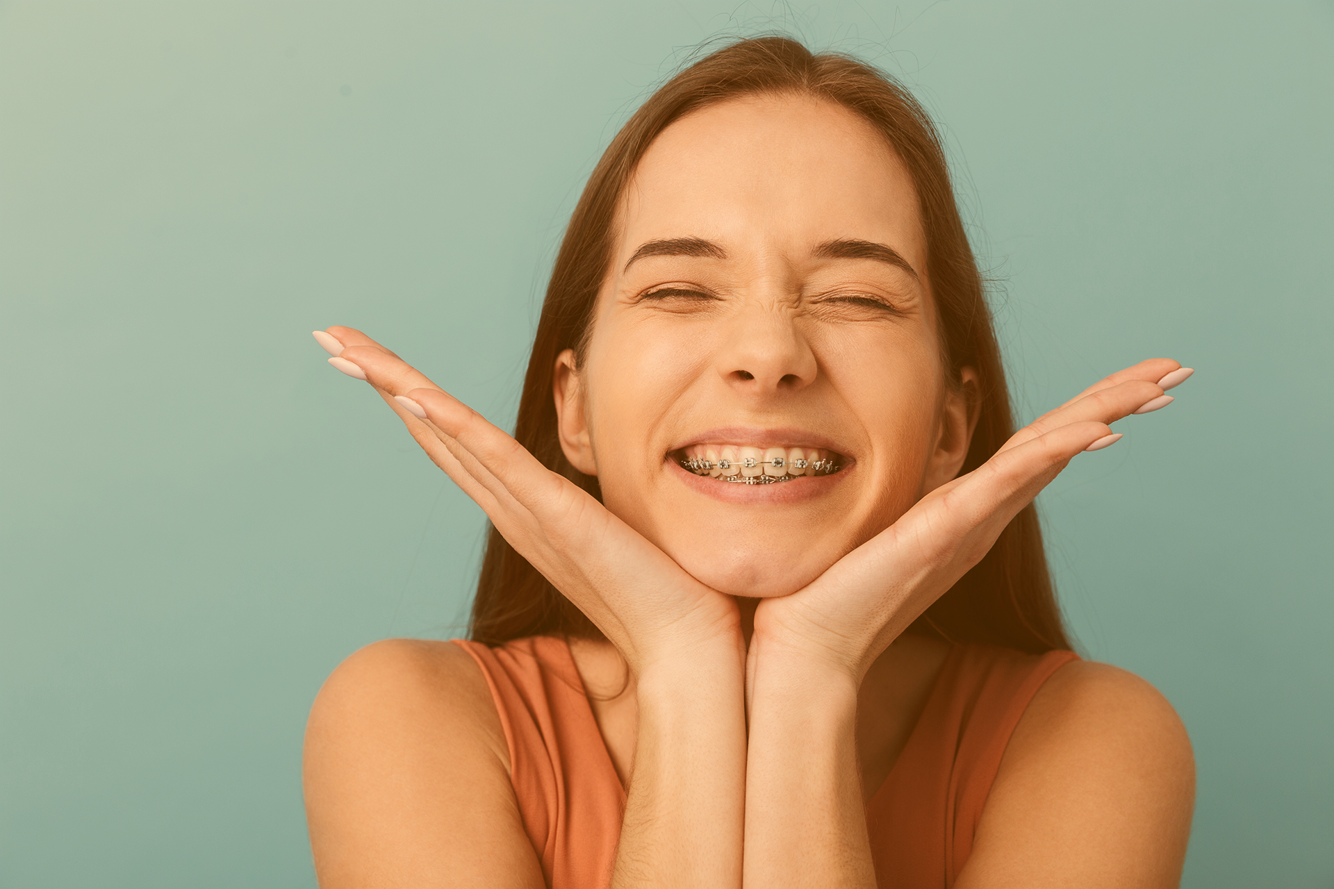 Brünette Jugendliche stützt ihr Kinn in ihre Handflächen und schenkt uns ein breites, mit einer Brackets-Zahnspange besetztes, Lächeln 