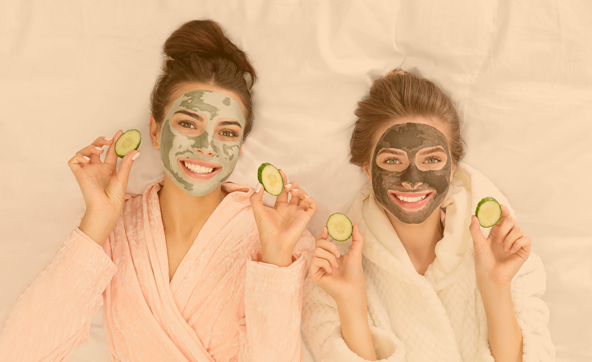 Zwei Freundinnen liegen im Bademantel und grüner Gesichtsmaske im Bett und halten je beidhändig Gurkenscheiben in den Händen