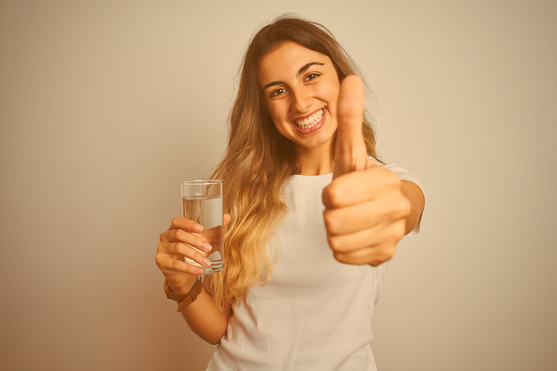 Junges, langhaariges Mädchen hält in ihrer einen Hand ein Glas Wasser und macht mit der anderen Hand das Daumen-hoch-Zeichen 