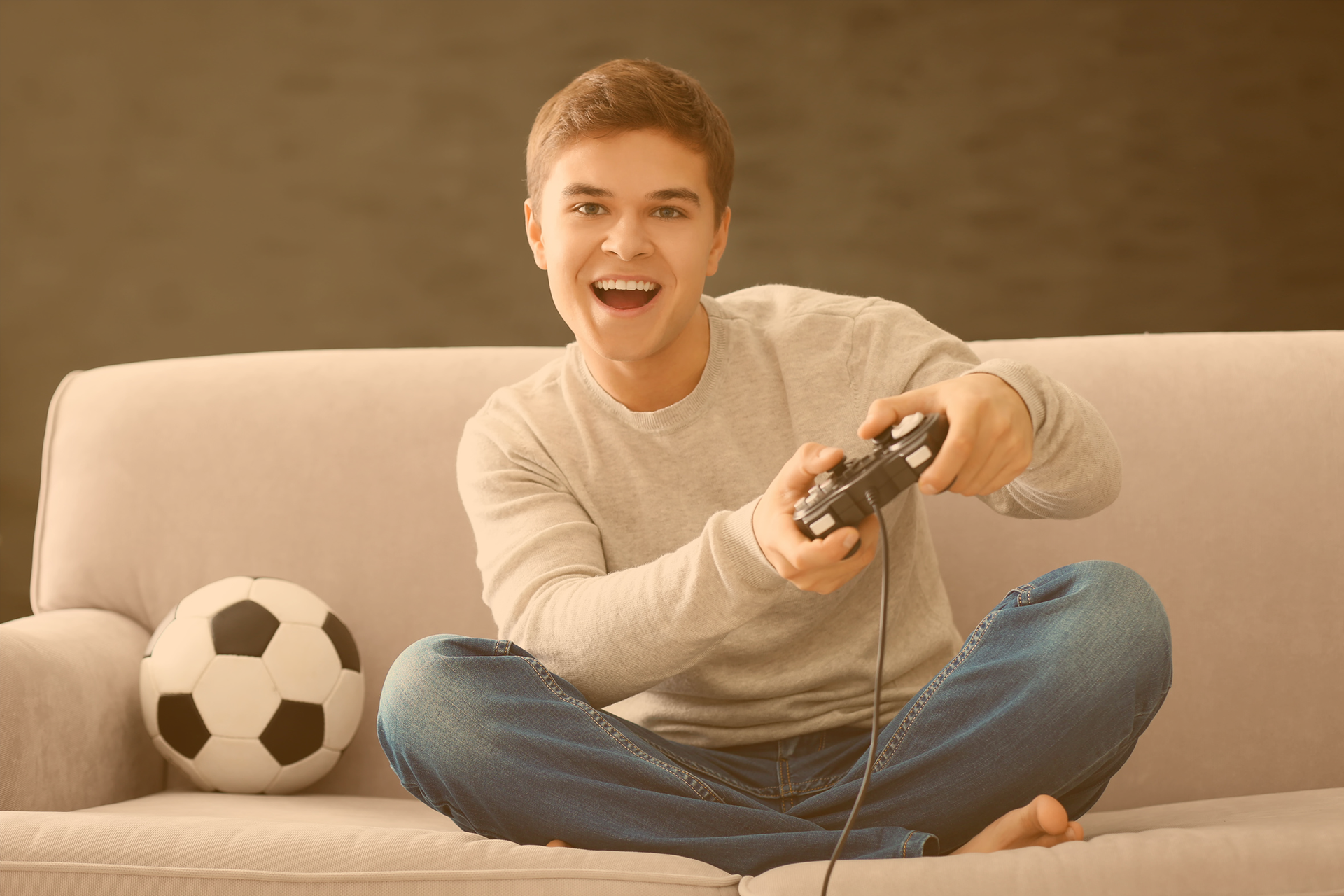 Teenager sitz im Schneidersitz mitfiebernd auf der Couch und hält eine Spieleconsole in seinen Händen, neben ihm liegt ein Fußball 
