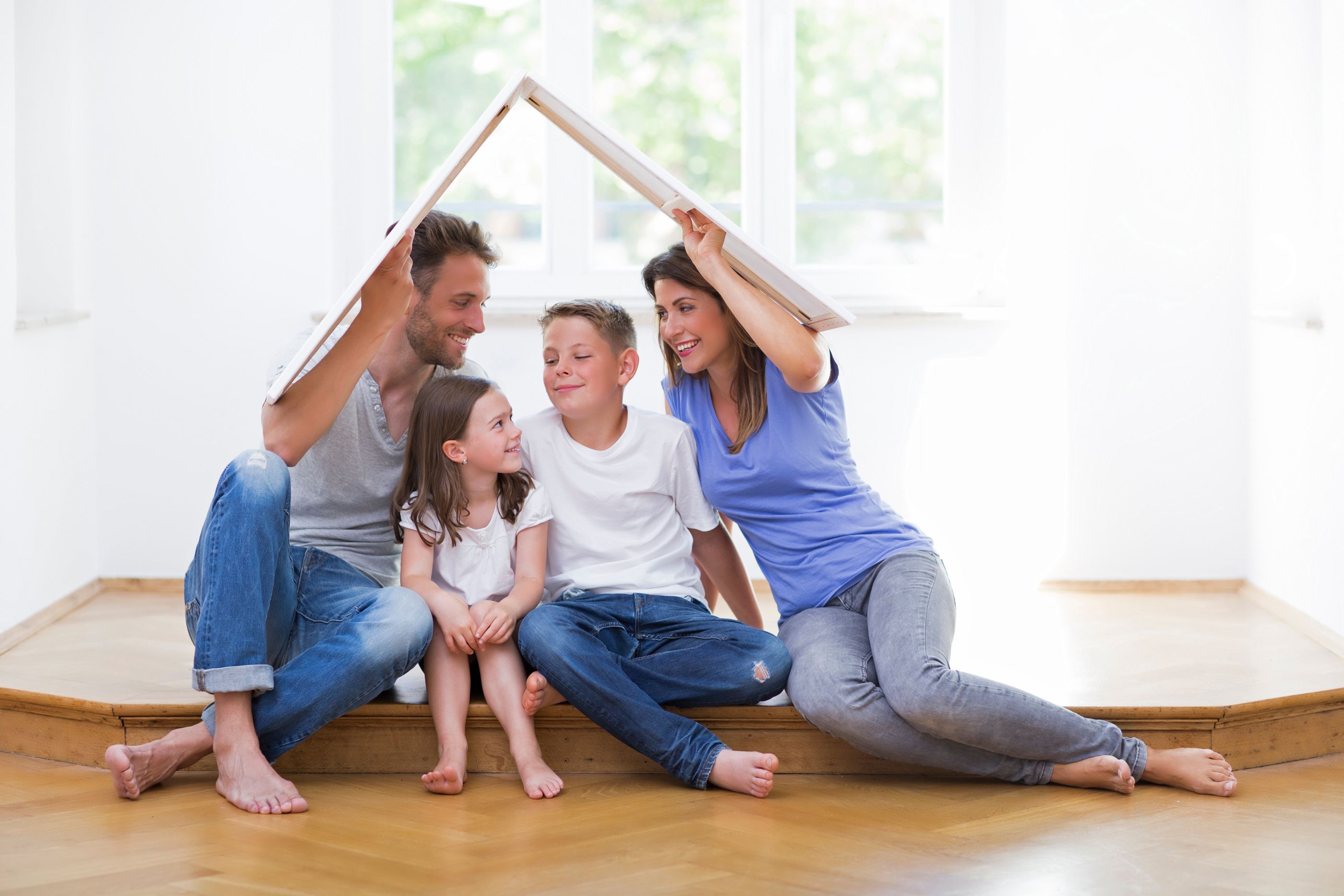 Familie sitzt glücklich in einer neuen Wohnung und hält sich symbolisch ein Pappdach über die Köpfe